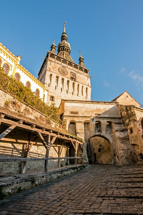 A cobblestone street leads through a tunnel under a large clock tower at sunrise