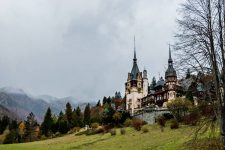 A white and brown Peles Castle on a green hillside, mountains in the background.