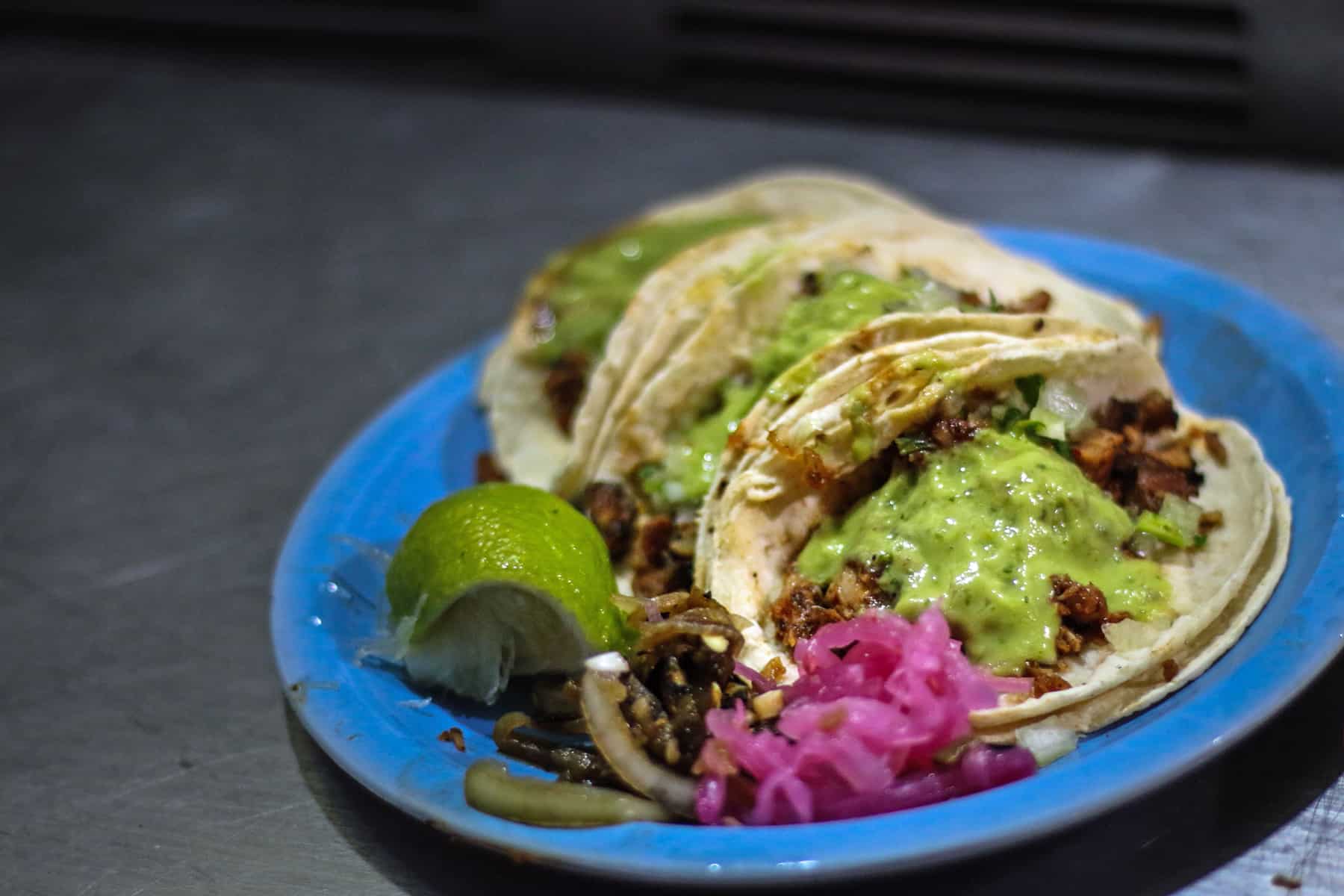 Blue plate with three tacos, a lime wedge, and red pickled onions. The best food in Oaxaca