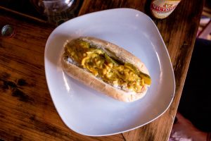 Plate with a hotdog in the centre, topped with pickles, onions and a yellow sauce.