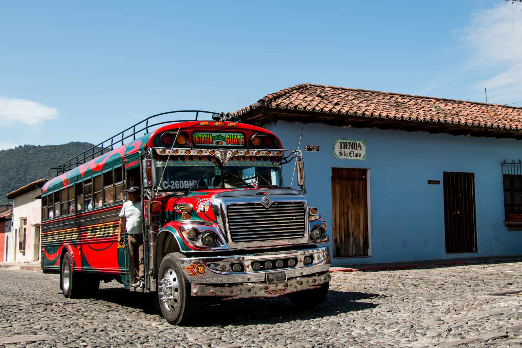 Red bus with flashy decals and stickers passing a blue building. This is one of the buses that can take you from Antigua to Panajachel