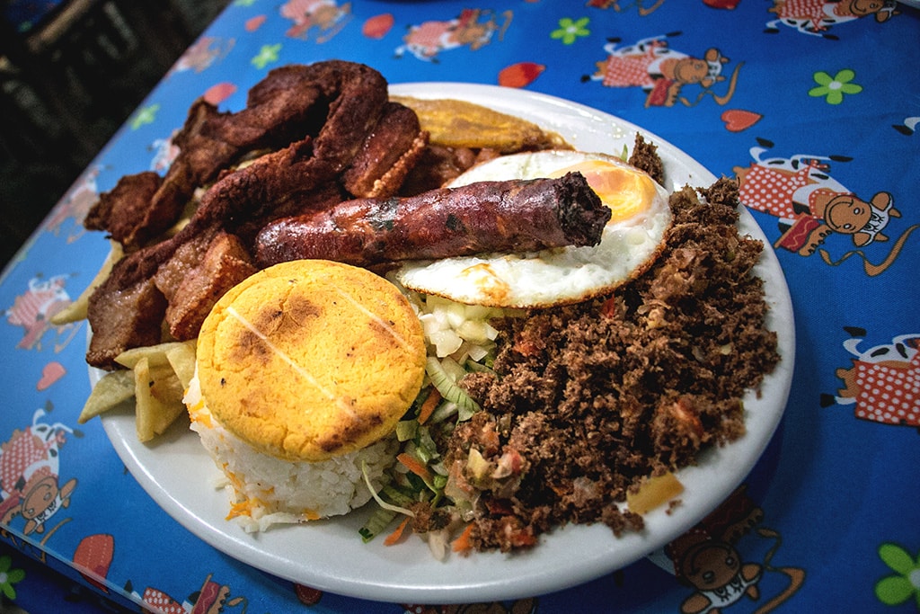White plate covered in a traditional Colombian dish featuring beans, sausage, bacon, eggs, potatoes and avocado.