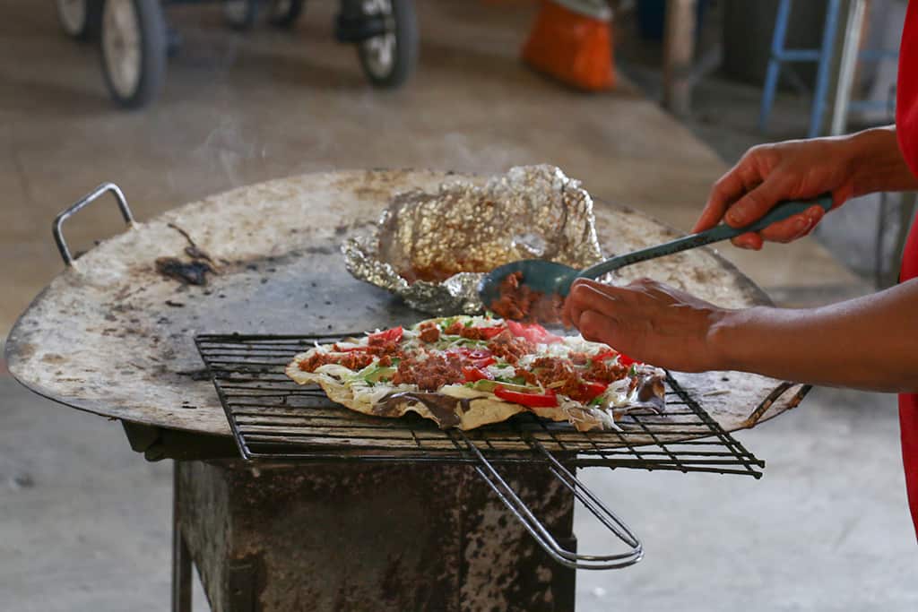 A woman placing toppings on a tortilla roasting over a charcoal grill.