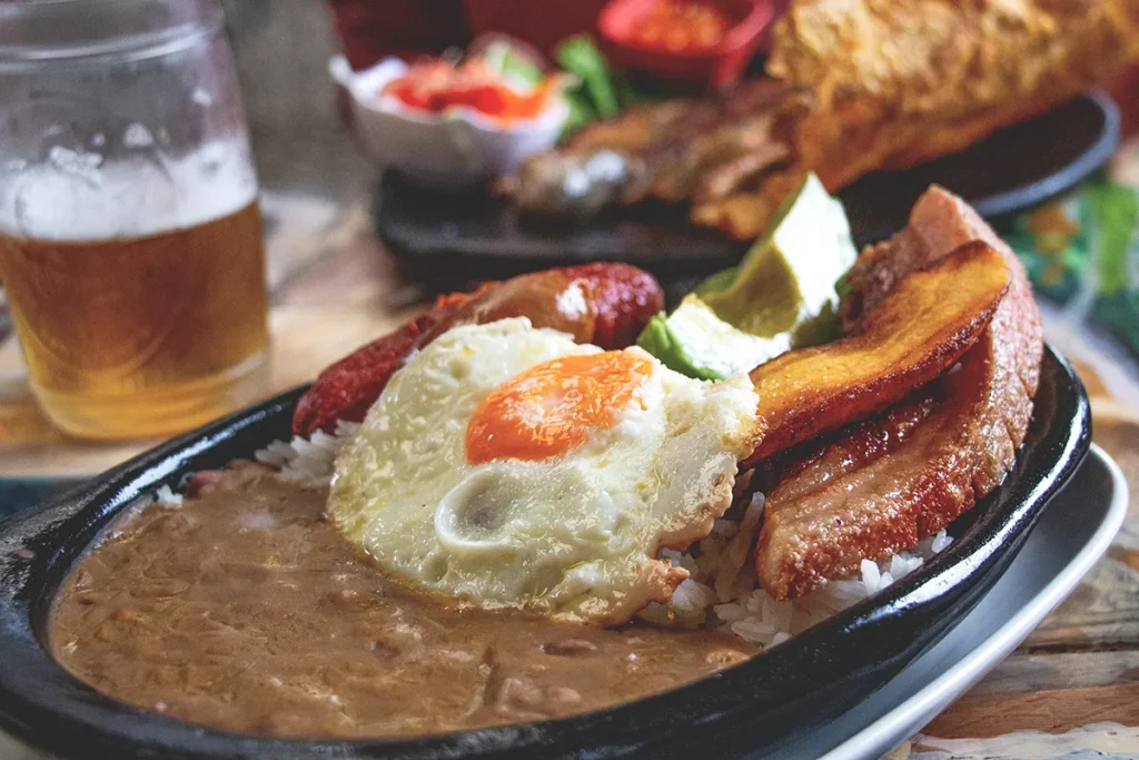 White plate covered in a traditional Colombian dish featuring beans, sausage, bacon, eggs, potatoes and avocado.