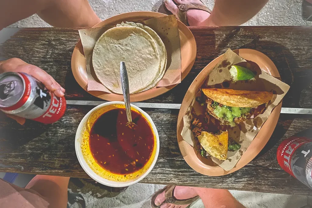 A wooden bench with a plate of tacos, a plate of tortillas and a bowl of red broth. Hands can be seen holding cans of beer.