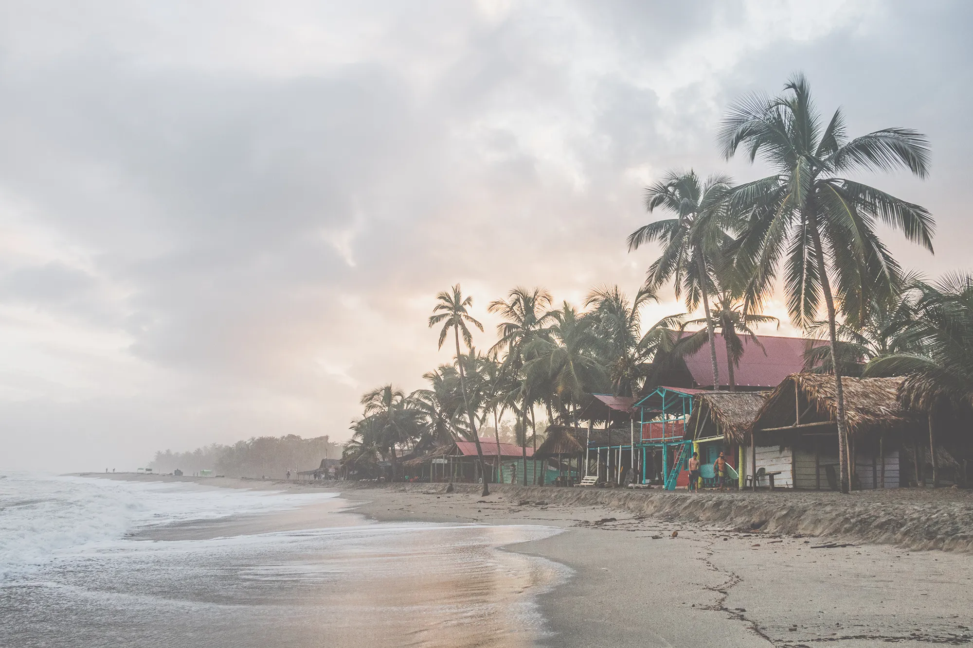 Image of a coastline at sunset with colourful wooden huts and palm trees along the sand.