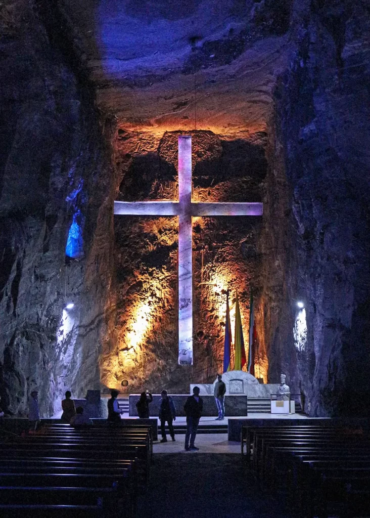 Dark cavern with purple and orange lights. A large cross hangs against the far wall with people standing beneath it.